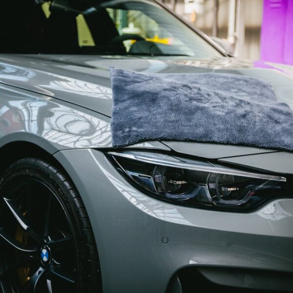 Gros plan sur l'avant d'une voiture grise, avec une serviette en microfibre Nanolex Allround Duo gracieusement posée sur le capot. La voiture est dotée de phares élégants et d'un logo de marque visible, avec un éclairage au plafond à l'intérieur se reflétant brillamment sur sa surface. - Tekalab