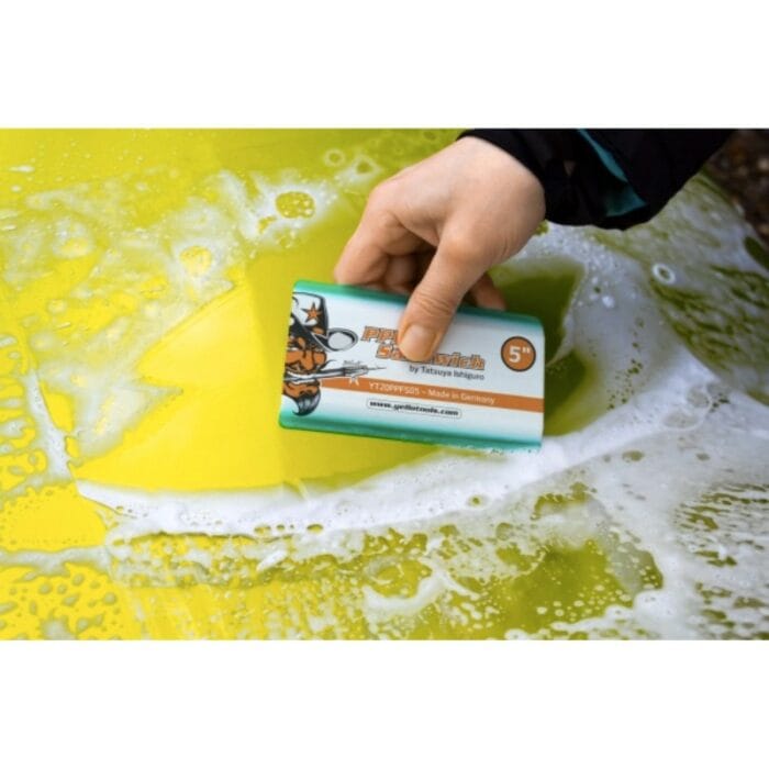 On voit la main d'une personne tenant un sandwich PPF, une raclette multicouche jaune et verte utilisée pour les films de protection de peinture, alors qu'elle nettoie une surface savonneuse, probablement une voiture. La raclette comporte une illustration et des coordonnées. - Tekalab