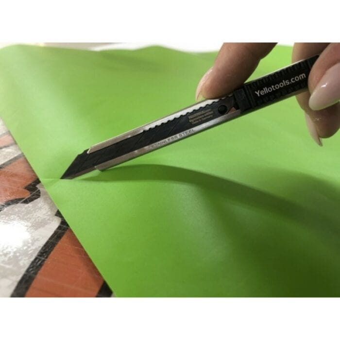 Une main tenant un couteau utilitaire BlackBlades doté de lames cassables extra-tranchantes coupe une feuille de papier vert. Le couteau noir, affichant la marque Yellotools sur le manche, démontre sa précision dans cette image en gros plan. - Tekalab