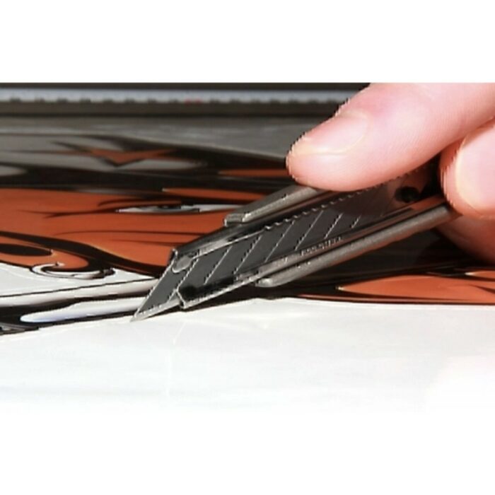 Gros plan d'une main maniant le YelloCut M Pro, un cutter professionnel pour les fabricants d'enseignes, coupant avec brio des matériaux imprimés aux motifs noirs et orange. L'accent est mis sur la lame, positionnée avec précision contre la surface à découper. - Tekalab