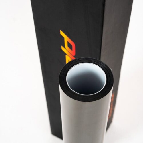 Une feuille de TPU FLEXISHIELD PPF WSH 8,0 mil laminée se trouve verticalement à côté dune boîte noire avec un lettrage coloré, capturé den haut pour mettre en valeur la forme cylindrique du vinyle sur un fond clair. - Tekalab