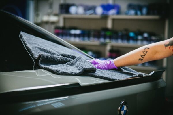 Une personne portant des gants violets utilise une serviette grise Nanolex Microfiber Allround Duo pour nettoyer la lunette arrière et le coffre d'une voiture. Le logo de la voiture est visible sur le coffre, avec des étagères garnies de diverses bouteilles en arrière-plan. - Tekalab