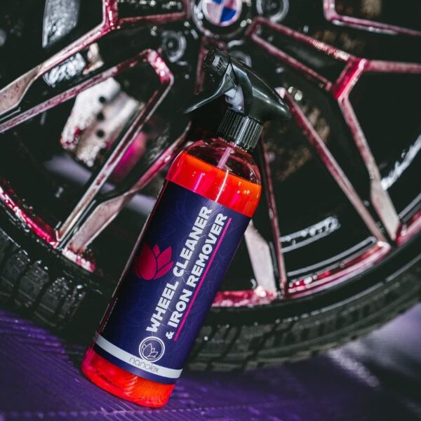 Une bouteille de nettoyant pour jantes et décapant pour fer Nanolex repose sur le sol près dune roue de voiture, en partie recouverte dun liquide rouge. Son étiquette présente un design violet accrocheur avec un texte blanc clair et rouge vif. - Tekalab