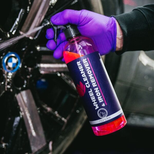 Une personne portant des gants violets tient un vaporisateur Nanolex Wheel Cleaner & Iron Remover avec un nettoyant rose vif devant une roue de voiture noire, avec un logo bleu et blanc. - Tekalab