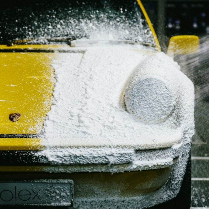 Une voiture de sport jaune brille de mille feux lorsqu'elle est traitée avec la mousse prélavée Nanolex, sa moitié avant étant recouverte d'une couche de mousse blanche. Les phares et la calandre avant de la voiture sont partiellement visibles, tandis que des gouttes d'eau scintillent sur sa surface lisse. - Tekalab