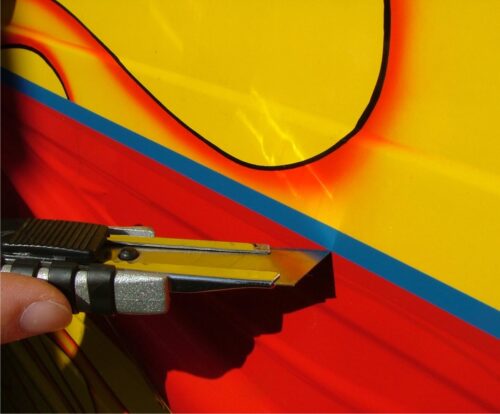 Gros plan d'une main utilisant habilement le cutter à ruban adhésif FineLine pour couper le long d'une ligne détaillée sur une surface aux couleurs vives. La surface est principalement jaune avec des accents rouges et orange, tandis que la lame est précisément inclinée pour une coupe précise. - Tekalab