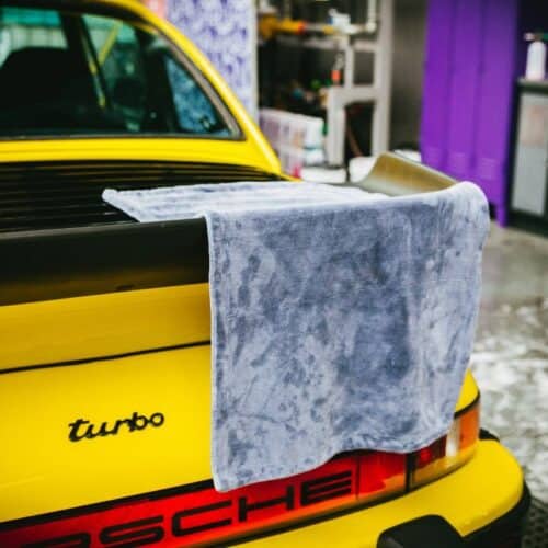 Une voiture de sport jaune équipée d'un turbo et des emblèmes Porsche à l'arrière est garée à l'intérieur. Drapée sur le spoiler arrière se trouve une serviette de séchage Nanolex Ultra. Le décor comprend une armoire violette ainsi qu'un éventail d'articles de garage. - Tekalab