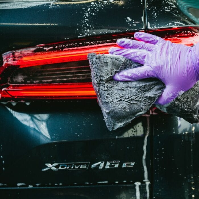 Enfilant une paire de gants violets, quelqu'un utilise un tampon de lavage Nanolex Ultra Plush pour nettoyer en profondeur le feu arrière d'un véhicule de couleur sombre enveloppé de mousse de savon. L'emblème du modèle xDrive 45e de la voiture brille de mille feux à côté du feu arrière fraîchement poli. - Tekalab