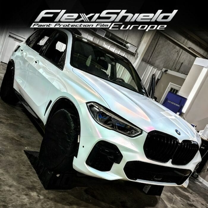 Un SUV BMW blanc est exposé dans une salle d'exposition, légèrement incliné vers la gauche. Les roues de la voiture sont dotées de housses de protection noires mises en valeur par le FLEXISHIELD Cosmétique PPF DM-01 Sparkling Pearl : Changez de couleur et protégez votre voiture. Au-dessus, un logo indique FlexiShield Paint Protection Film Europe, ajoutant à l'atmosphère moderne et industrielle. - Tekalab