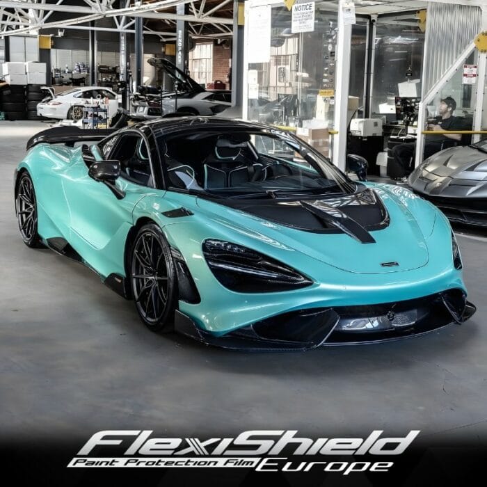 Une élégante voiture de sport turquoise aux accents noirs, protégée par le FLEXISHIELD Cosmétique PPF HG-07 MINT FRESH, est garée dans un garage spacieux. La voiture présente un design aérodynamique moderne, avec dautres véhicules et équipements automobiles visibles en arrière-plan. - Tekalab