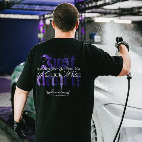 Un homme portant un t-shirt noir GreenX avec le texte décoratif « Violet : Just Clean It » lave au jet deau une voiture, qui apparaît mouillée et brillante en arrière-plan. - Tekalab