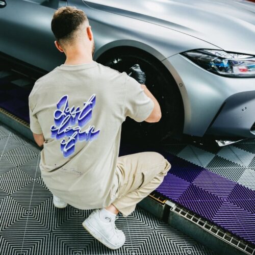 Une personne portant un T-shirt beige GreenX avec linscription « Just clean it » en violet est accroupie à côté dune voiture argentée élégante, en train de polir ses roues. Ils portent un pantalon assorti et des baskets blanches sur un sol à motifs déléments géométriques violets et gris. - Tekalab