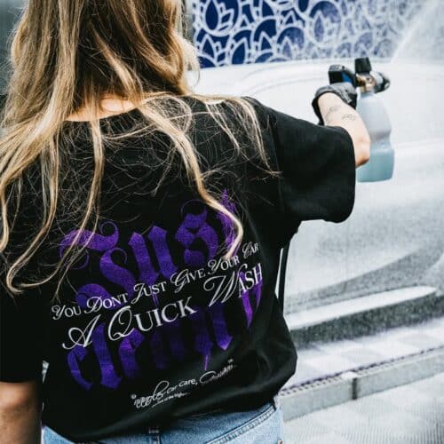 Une personne aux cheveux longs portant un T-shirt noir GreenX avec le texte « Just clean it » en violet utilise un pistolet pulvérisateur pour laver une voiture. Larrière-plan montre un véhicule partiellement lavé. - Tekalab