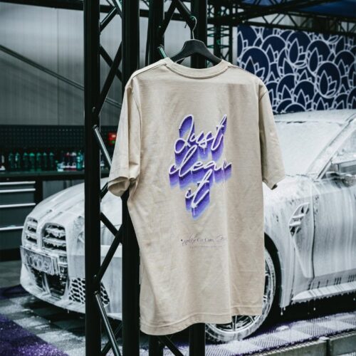 Un T-shirt GreenX beige avec le texte violet « Just clean it » est accroché dans un garage. Derrière lui, une voiture blanche savonneuse est entourée de fournitures automobiles. - Tekalab