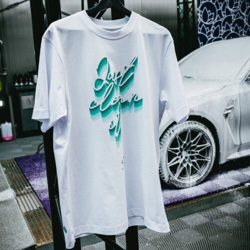 Un T-shirt GreenX noir/violet avec la phrase « Just clean it » est accroché à une balustrade. En arrière-plan, une voiture blanche est recouverte de savon, capturant parfaitement lessence dune scène de lavage de voiture animée. - Tekalab