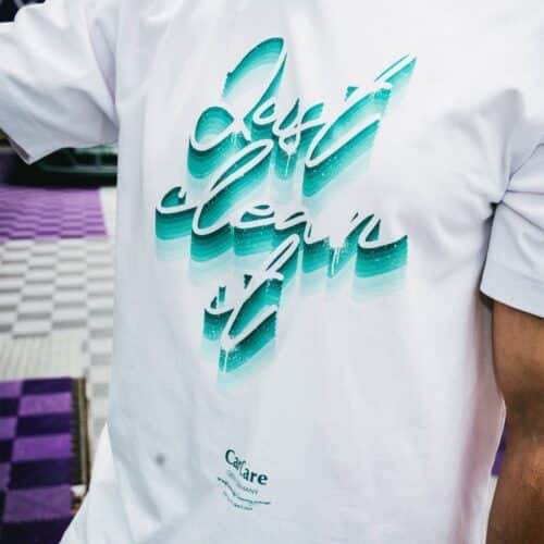 Un gros plan montre quelquun portant le T-shirt GreenX Noir/Violet, présentant « Just clean it » dans un dégradé bleu-vert vibrant sur un sol à carreaux violet-gris, accentuant lesthétique Violet. - Tekalab