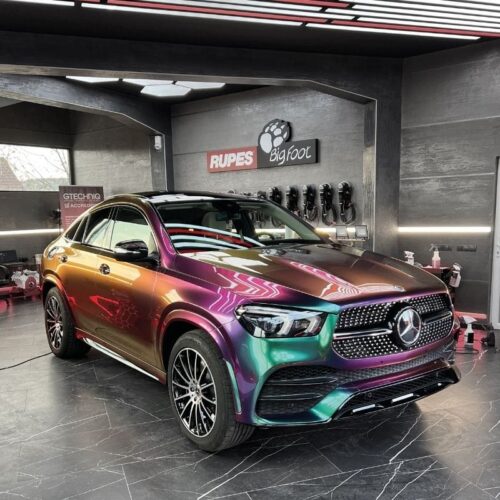 Un SUV Mercedes-Benz avec une finition caméléon vibrante de la palette FLEXISHIELD Cosmétique PPF TP-04 Moon Beam 2.0 dans des teintes violettes, vertes et dorées est garé à lintérieur dun garage de détail moderne doté de murs noirs élégants et déquipements automobiles. - Tekalab