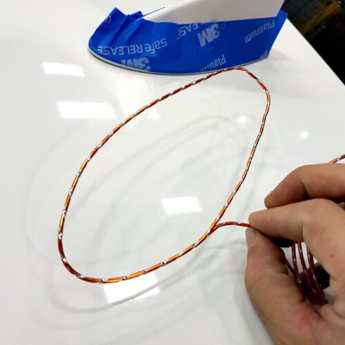 Une main tient une boucle flexible de fil fin, brillant et torsadé orange WrapBend-It | File de coupe sur un fond blanc. Un rouleau de ruban adhésif bleu 3M partiellement visible est en arrière-plan. - Tekalab