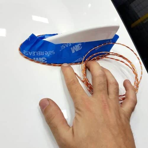Une main ajuste une antenne WrapBend-It avec une base blanche, fixée par du ruban adhésif bleu 3M sur une surface blanche. Un fil spiralé orange et blanc sétend gracieusement, assurant un positionnement optimal pour une connectivité transparente. - Tekalab