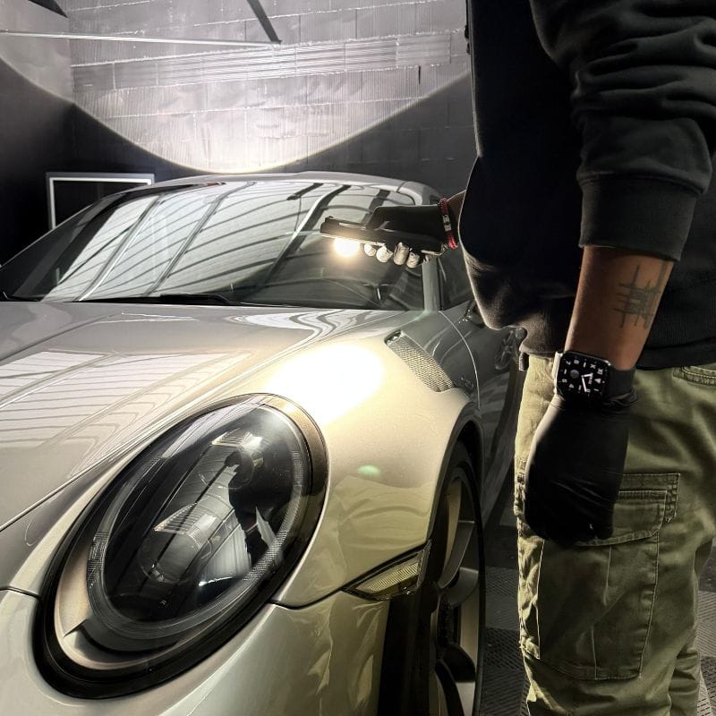 Une personne portant des gants inspecte la surface dune voiture argentée élégante avec une lampe portative, mettant en valeur sa finition brillante. Malgré lextérieur impressionnant, on craint quune mauvaise odeur de voiture ne persiste à lintérieur. Le décor semble être celui dun atelier de détail ou de réparation automobile, se concentrant sur la zone avant. - Tekalab